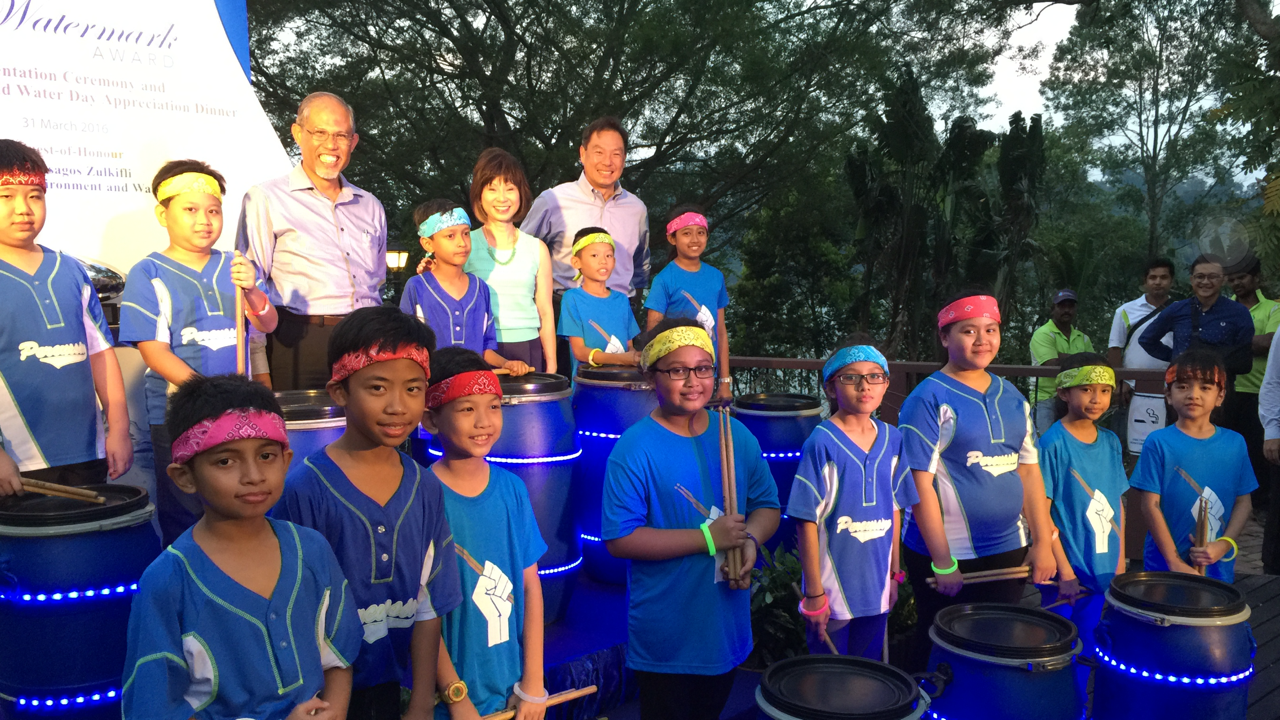 Children in blue uniforms with colorful headbands pose with drumsticks beside lit drums at a Watermark Awards event.